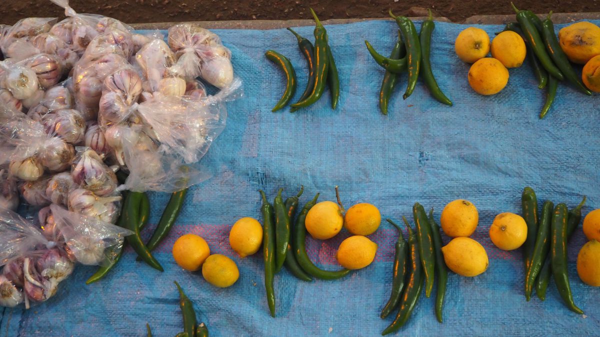 Auf dem Marktplatz wurde ein hellblauer Sack aus Kunststoffgewebe ausgebreitet, darauf liegen portionsweise jeweils 3 leuchtend gelbe Zitronen, 4 grüne Pepperoni sowohl auch in transparenten Plastikbeuteln abgepackte weiß-violette Knoblauchknollen.