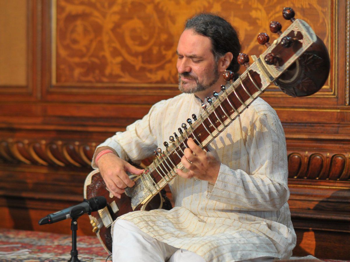 Auf einem orientalischen Teppich sitztbarfuß und im offenen Schneidersitz ein Musiker und spielt auf der Sitar, einer Langhalslaute.