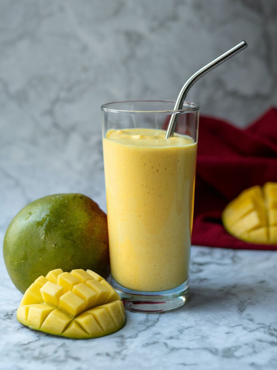 In einem Glas mit Edelstahltrinkhalm das erfrischend gelbe Getränk aus Manosaft und Joghurt, daneben eine ganze Mangofrucht sowie eine kunstvoll würfelig aufgefächerte Hälfte.