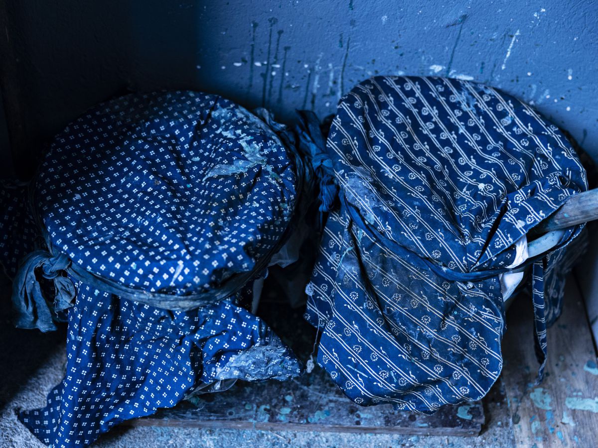 Das Bild ist ganz von dunklem Blau dominiert: Zwei Farbkübel, aus einem ragt ein hölzener Stiel, beide sind mit Tüchern bedeckt und zugebunden. Die Stoffe tragen weiße, feine Muster auf intensivem blauen Grund. Der Boden und die Wand sind von blauen Farbs