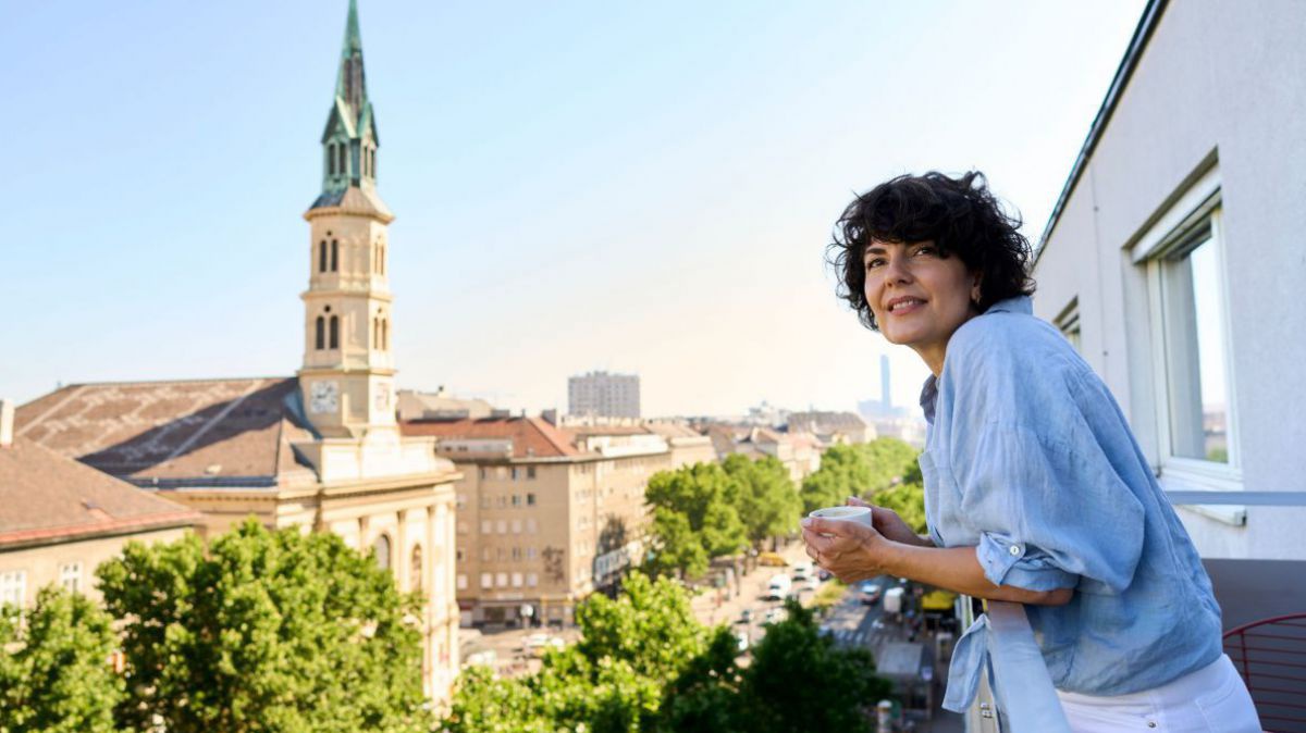 Frau steht mit Kaffeehäferl in der Hand am Balkon und genießt den Weitblick.