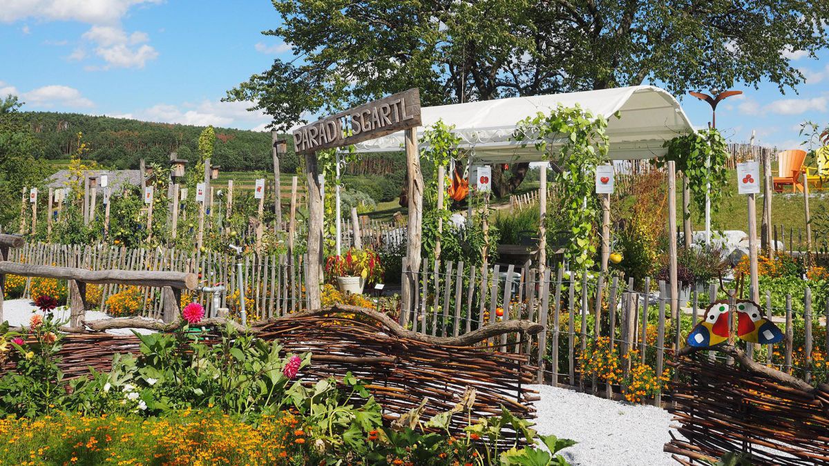 Weidenzaun, darin ein Tor mit einer Holztafel über dem Einhang auf der Paradiesgartl steht, dahinter eine üppig blühende Gartenlandschaft.