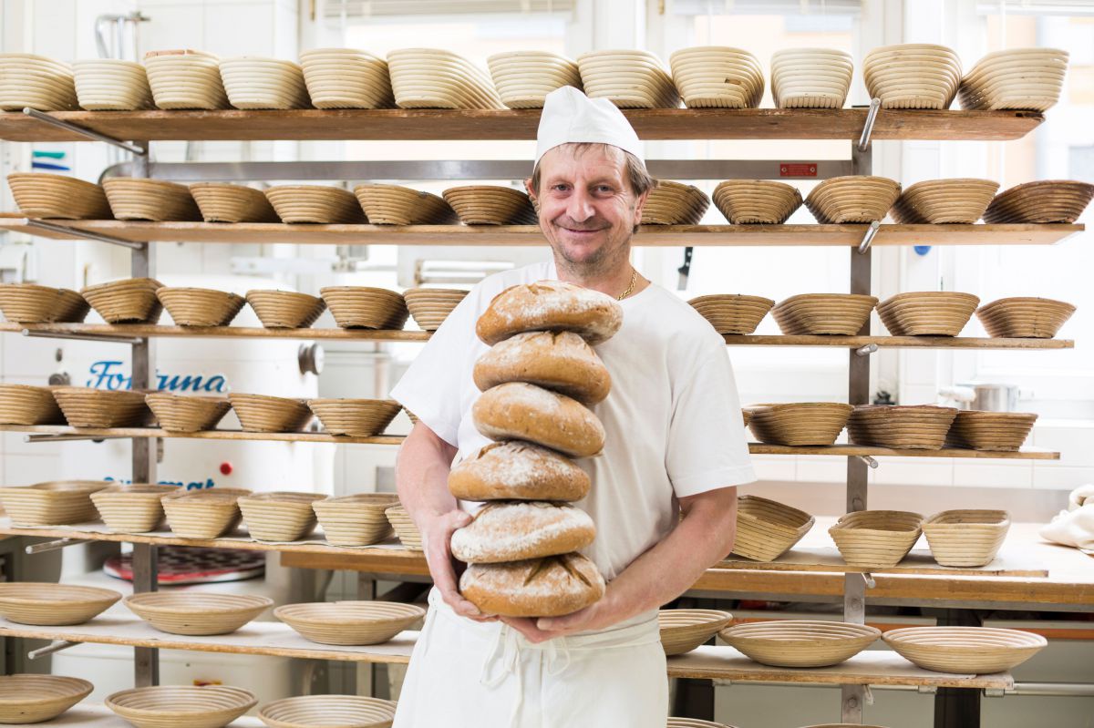 Bäcker mit weißer Schürze und Mütze trägt 6 übereinander gestapelte Brotlaibe, dahinter ein Regal mit Brotkörben. 