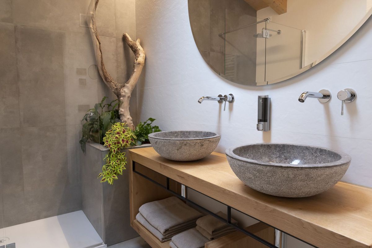 Waschtisch im Badezimnmer mit zwei runden Waschbecken aus grauem Stein. In der Ecke ein rechteckiger, steinerner Trog mit einem Stück Holt und grünen Pflanzen.