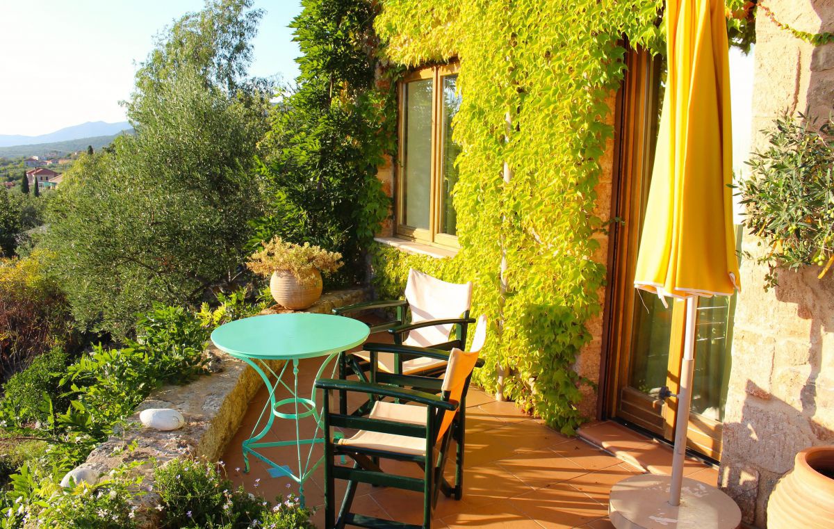 Mit Pflanzen bewachsene Hausfront, davo eine kleine Terase mit einem Tisch und zwei Stühlen.