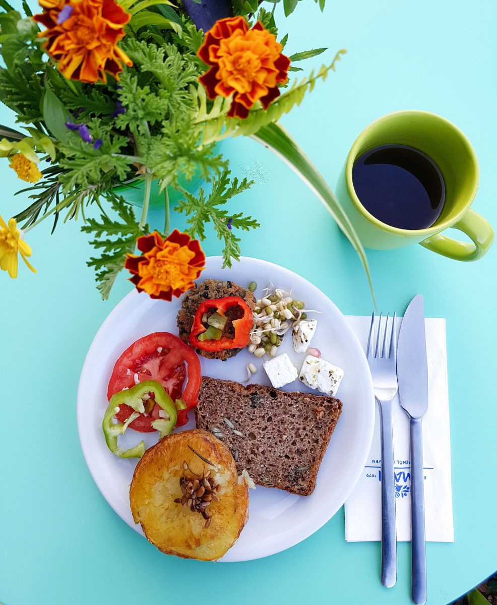 Teller mit Gemüse, Feta und Brot, eine Tasse Kaffee und ein Strauß Blumen auf einem Tisch von oben fotografiert.