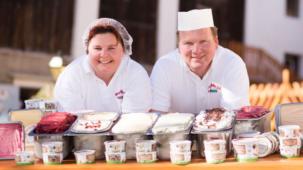 Petra und Bernd Pobaschnig haben beide ein weißes Gewand an. Sie lehnen mit den Armen auf einem Tisch und vor ihnen sind jede Menge Eissorten aufgebaut.