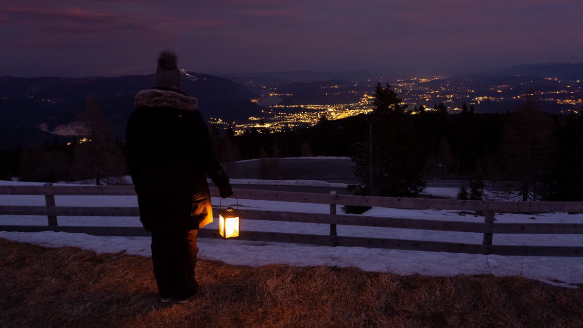 Frau mit einer Laterne steht in der Nacht auf einem Berghang hinter einem Lattenzaun und blickt auf die Lichter einer Stadt.