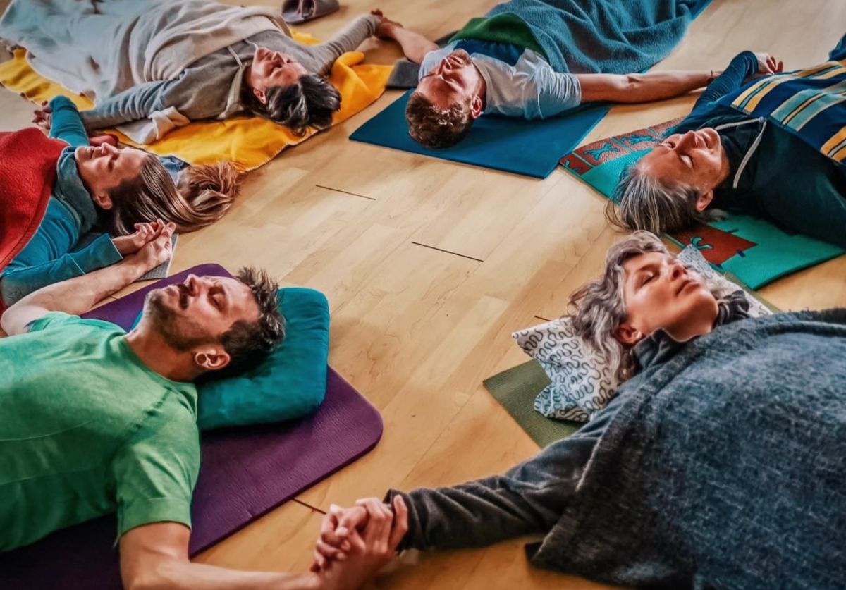 Gruppe von 6 Personen liegt in einem Raum im Kreis am Boden, alle halten sich an den Händen und üben die bewusste Atmung.