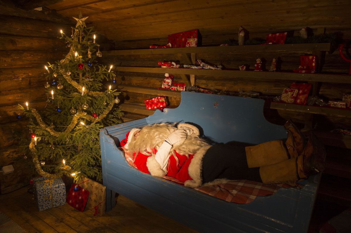 Weihnachtsmann liegt in einem Holzbett in einer Holzhütte, dahinter ein Christbaum, darunter und an der Wand ein Regal mit Packerl.