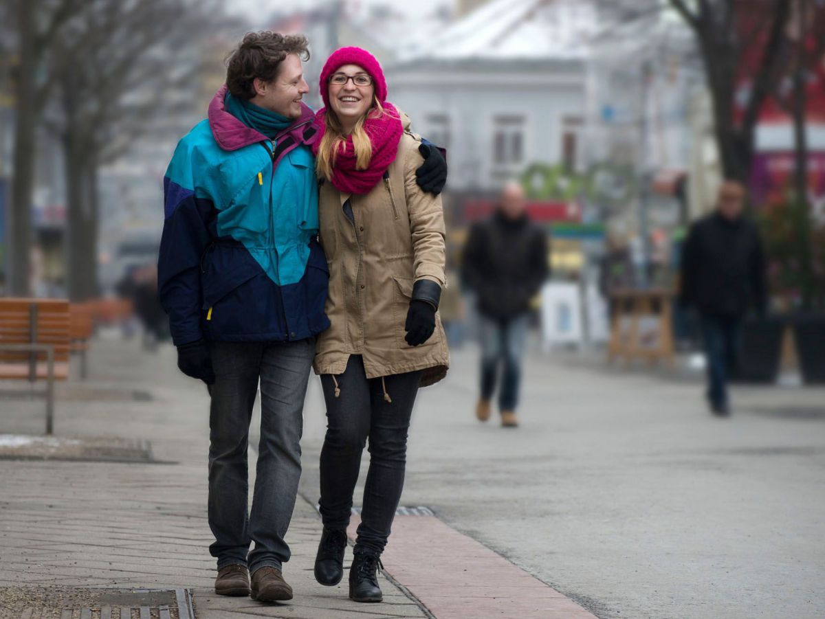 Ein Paar spaziert durch ein Stadtzentrum, sie trägt eine Haube und einen dicken Schal, beide warme Jacken und Handschuhe.