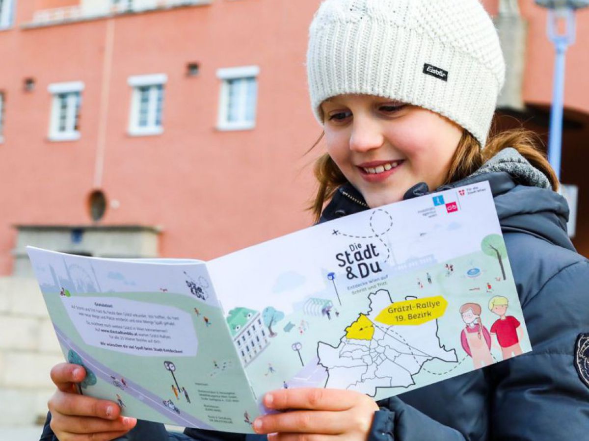 Ein Mädchen im Schulalter steht auf einem Platz in Wien und erkundet die Gegend mit einem Rätselheft von 