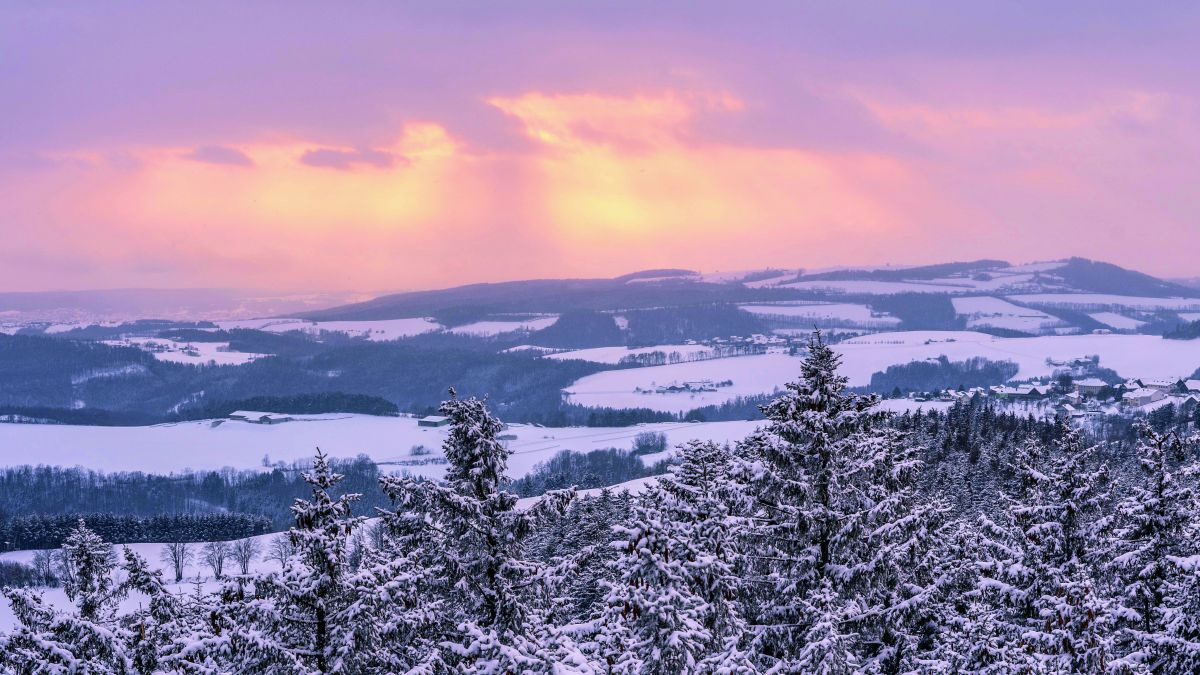 Orangeroter Himmel über leicht hügeliger und bewaldeter L
Winterlandschaft.