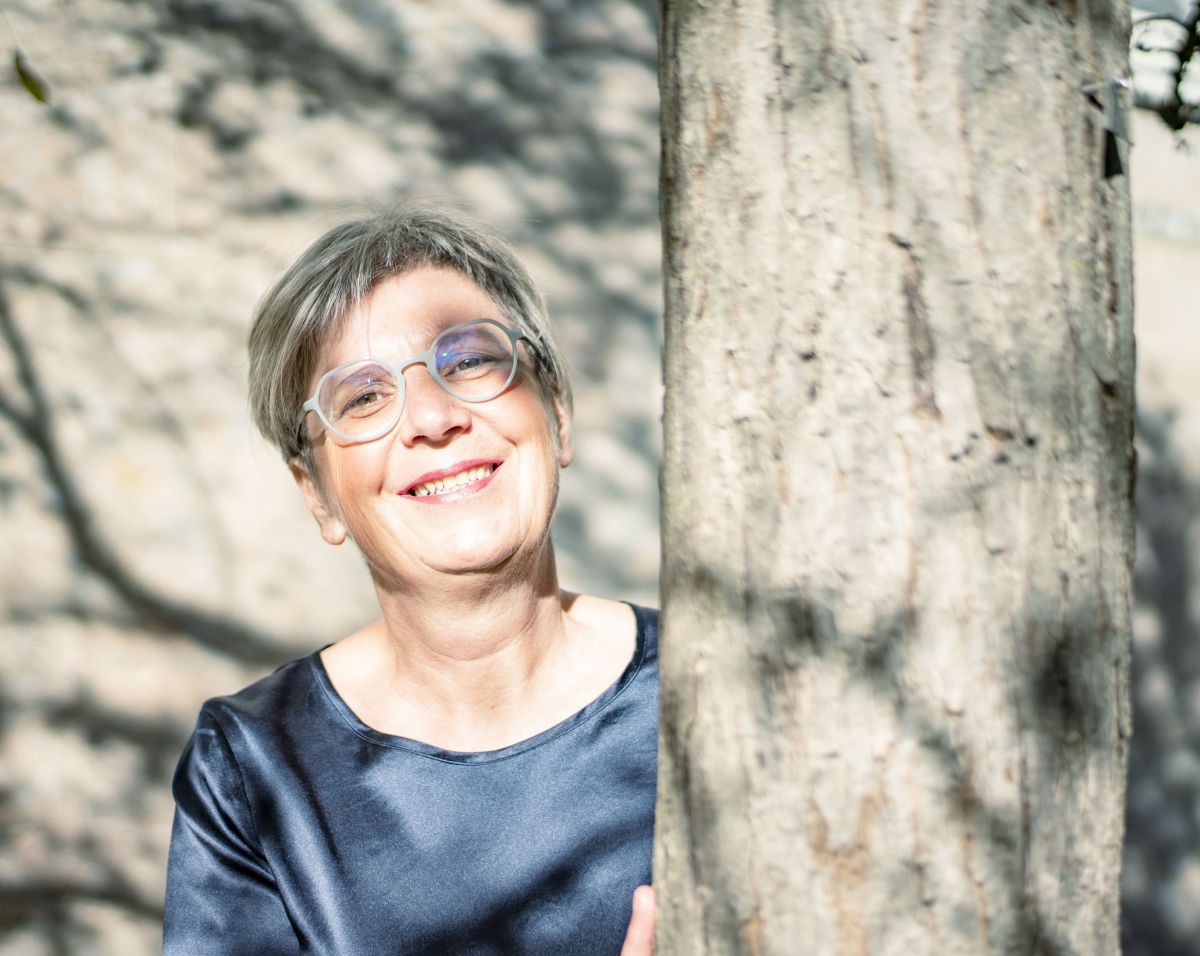 Portraitfoto von Klaudia Bestle, graue kurte Haare mit Brille, sie lehnt an einem Baum.