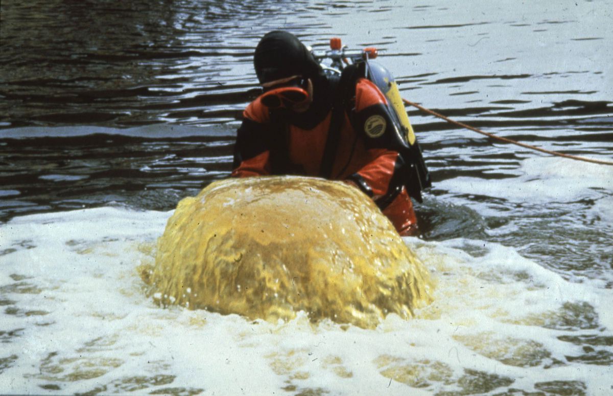 Mann im Taucheranzug im Wasser stehend, vor ihm sprudelt eine gelbe Flüssigkeit.
