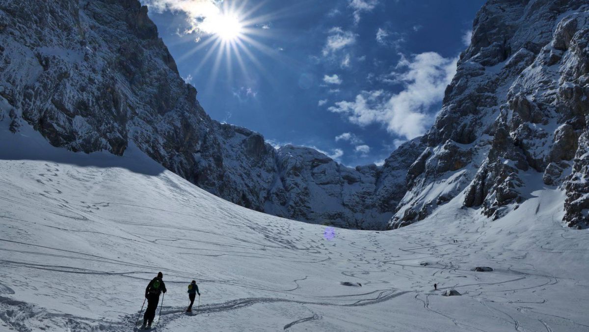 Zwischen den Felswänden blitzt eine strahlende Sonne auf blauem Himmel hindurch, die Skitourengeher ziehen ihre Spuren durch den Tiefschnee.