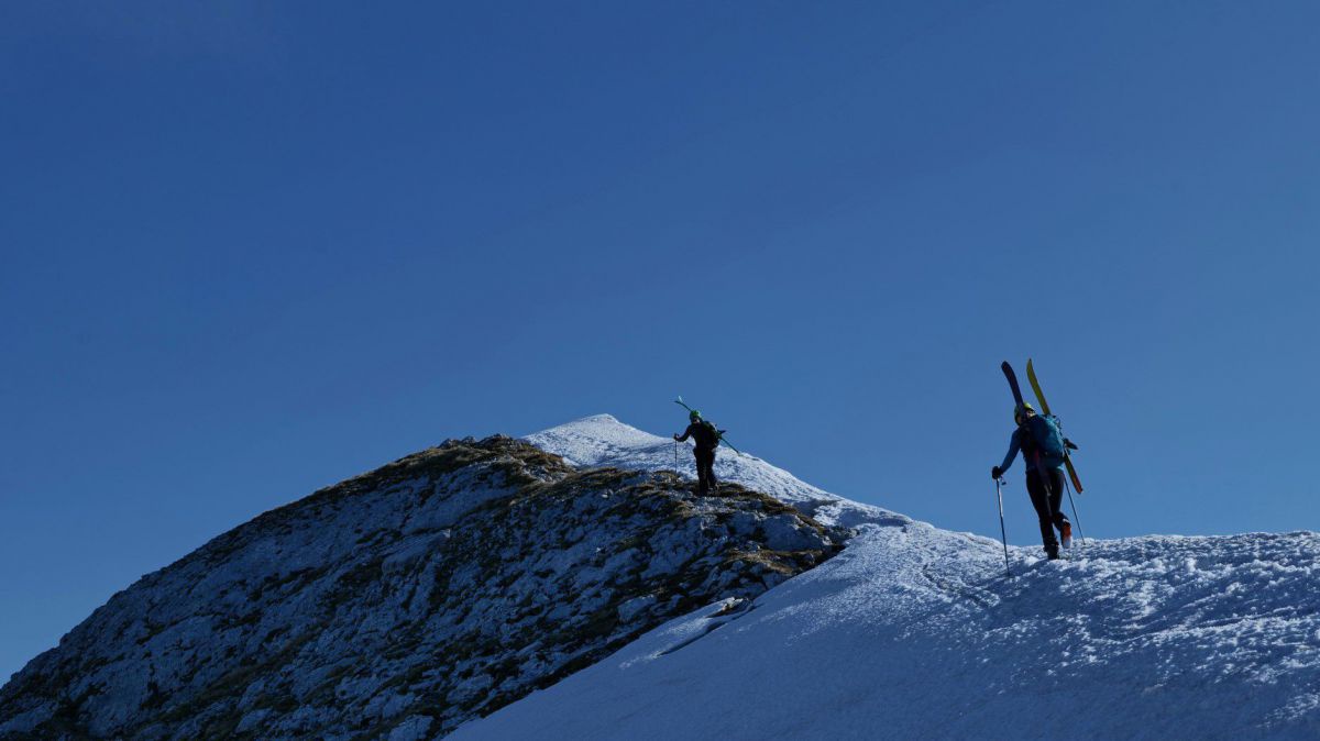 Zwei Tourengeher, die Ski auf den Rücken geschnallt, wandern über einen Kamm, der großteils von Schnee bedeckt ist, einem tiefblauen Himmel entgegen.