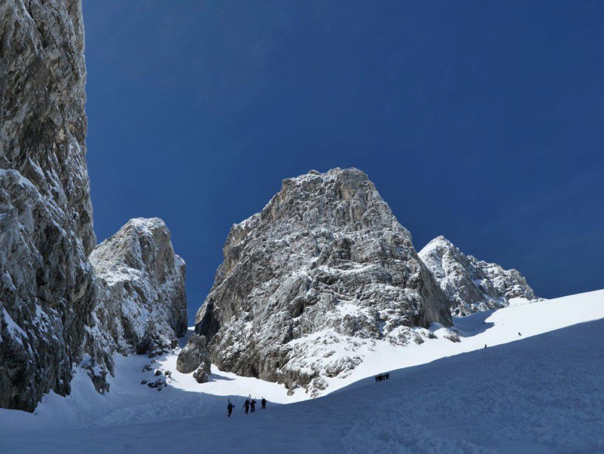Der Felsen im zentrum des Fotos wird won der Sonne beschienen, während die Skitourengeher winzig in der schneebedeckten Ebene unter dem Schatten einer mächtigen Felswand wandern.