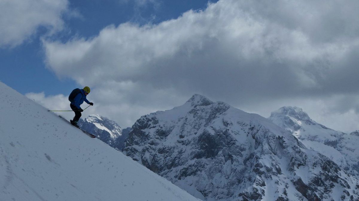 Ein Skifahrer bei der Abfahrt über einen steilen Hang, dahinter wolkenüberhangene niedrigere Berggipfel.