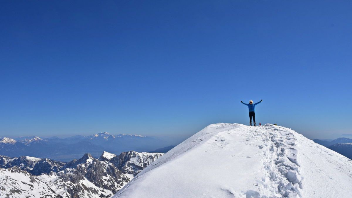 Eine Tourengeherin steht mit ausgebreiteten Armen auf dem tiefverschneiten Gipfel.