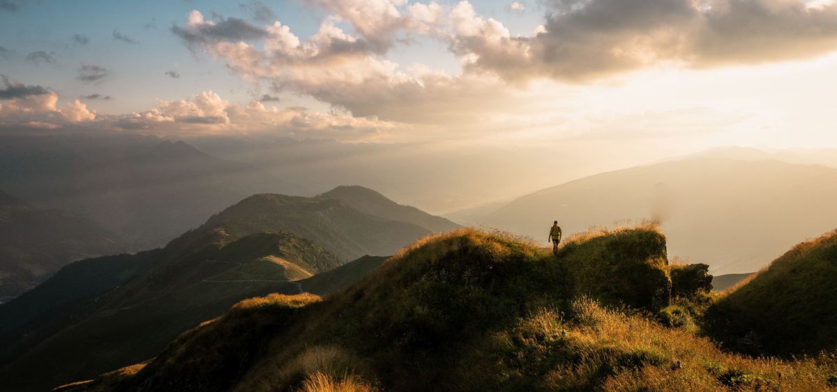 Wanderer auf einem grasbedeckten Höhenweg. Die Sonne blitzt hinter einigen Wolken hervor.