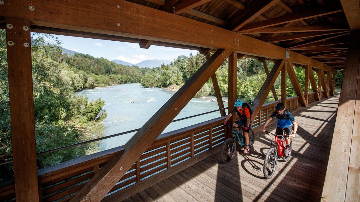 Zwei Radfahrer überqueren die Isel auf einer Holzbrücke.