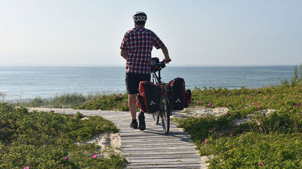 Ein Radfahrer schiebt sein bepacktes Fahrrad über einen mit Holzlatten befestigten Weg an der Küste. Im Hintergrund das Meer.
