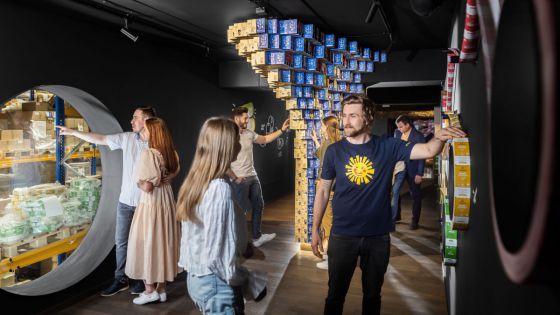 Ausstellung in einem dunklen Raum. Sonnentor-Mitarbeiter mit gelber Sonne auf blauem T-Shirt führt Besucher*innen durch die Schau.