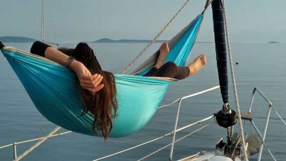 Eine langhaarige Person liegt in einer türkisblauen Hängematte auf einem Segelboot.