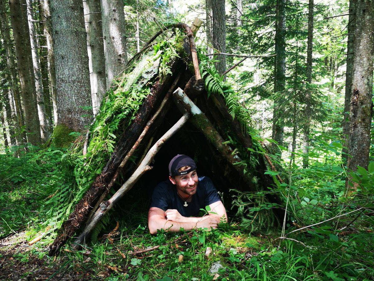 Ein Mann liegt unter seinem selbstgebauten Zelt aus Ästen, Farnen und Moosen in einem Wald.