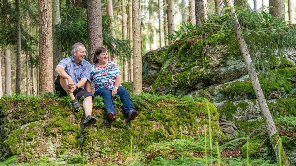 Ein älteres Paar sitzt auf einem stark bemoosten Felsen in einem Nadelwald beim 