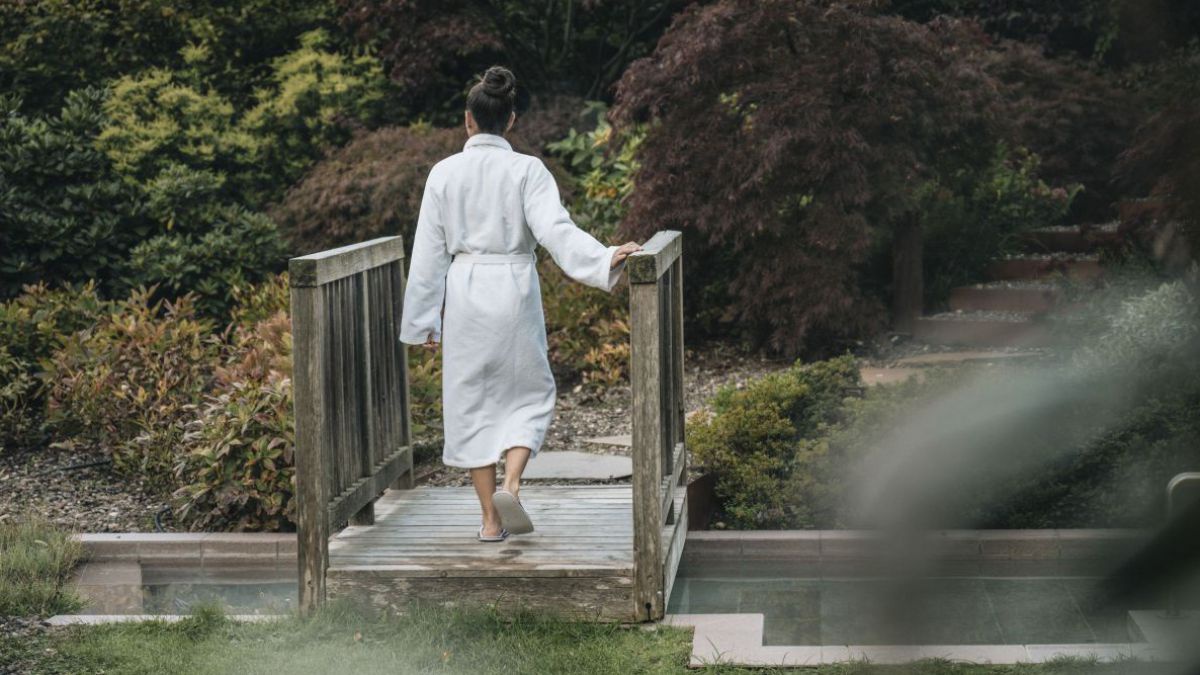 Eine Frau im weißen Bademantel schreitet über eine kleine Holzbrücke im Garten des Biorefugium Theiner´s Garten.