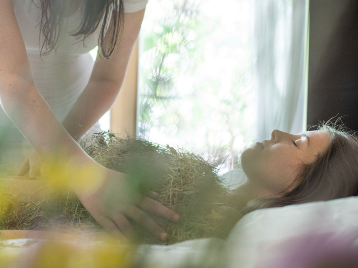 Eine Frau liegt mit geschlossenen Augen auf einer Therapieliege und erhält eine Wellnessbehandlung mit Heublumen von einer weiblichen Therapeutin.