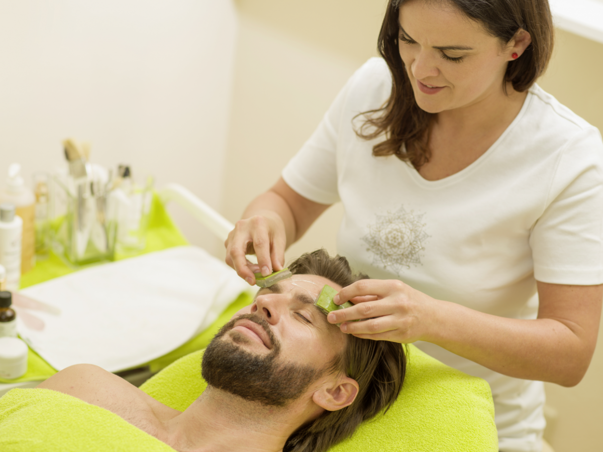 Ein bärtiger Mann liegt auf einer hellgrünen Therapieliege und erhält von einer weißgekleideten Frau mit dunklem langen Haar eine Gesichtsbehandlung mit Aloe Vera. Auf einem Beistellwagen stehen Fläschchen, Dosen und Pinsel.