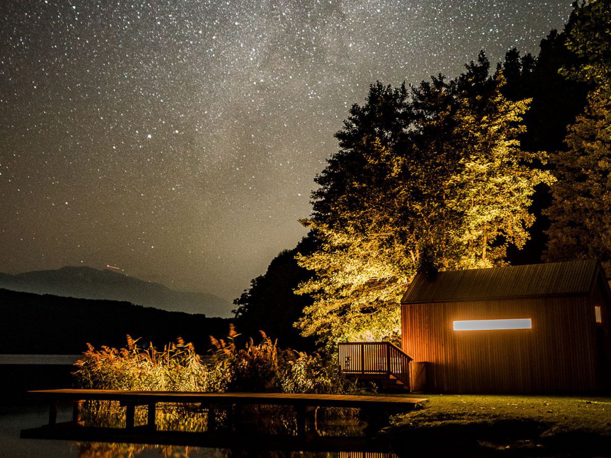 Ein Häuschen aus Holz, dezent beleuchtet, am bewaldeten Ufer des Millstätter Sees, ein hölzerner Steg führt durch Schilf zum Wasser, der Nachthimmel im Hintergrund ist von unzähligen Sternen übersät.