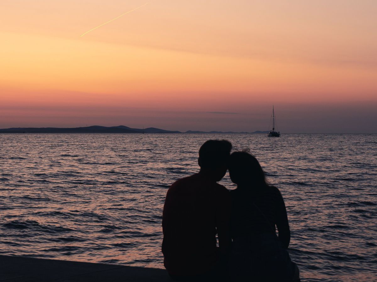 Ein Paar sitzt nach Sonnenuntergang am Strand, auf der leicht bewegten Wasserfläche des Meeres sieht man in einiger Entfernung ein Segelboot.