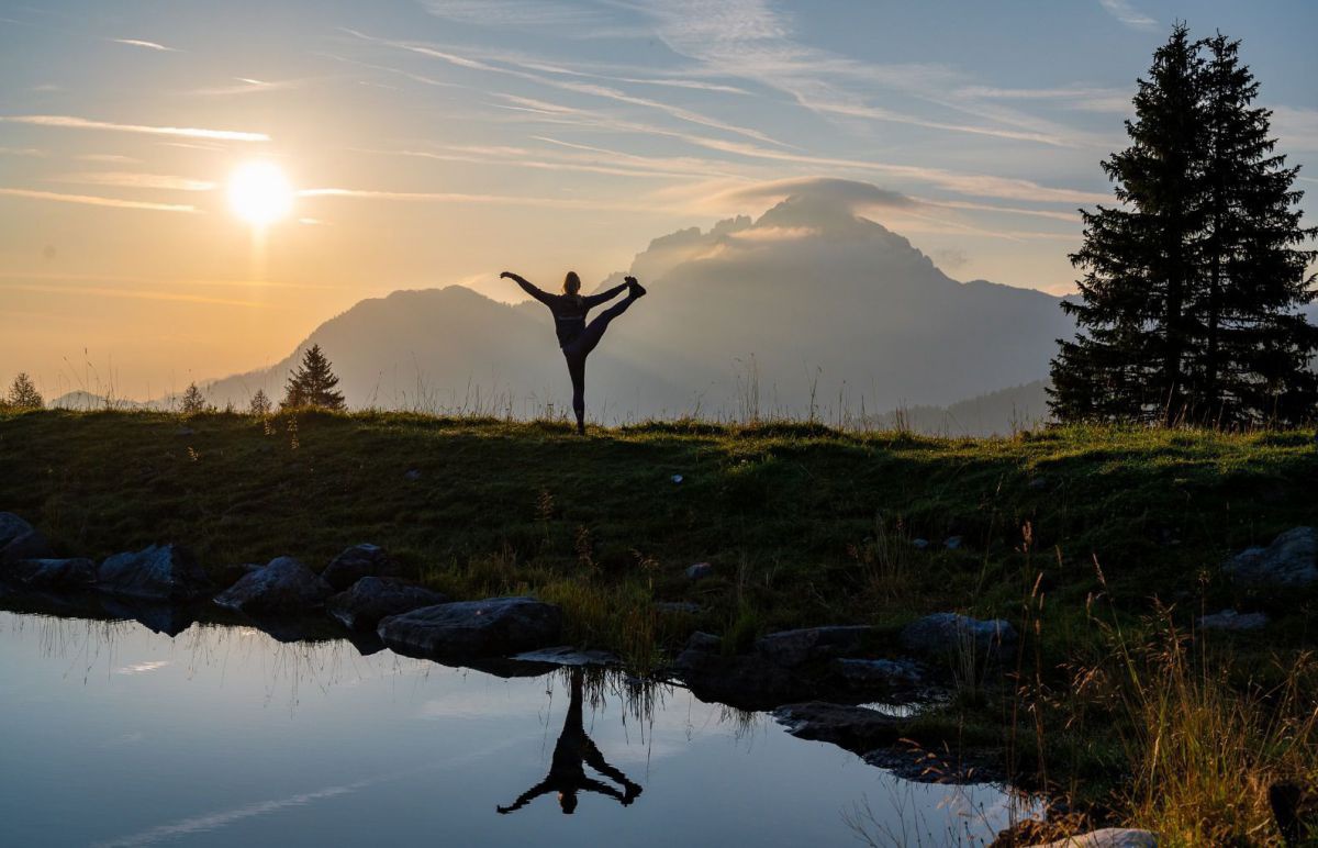 Person beim Yoga, im Vordergrund ein See, dahinter geht die Sonne über den Bergen auf.