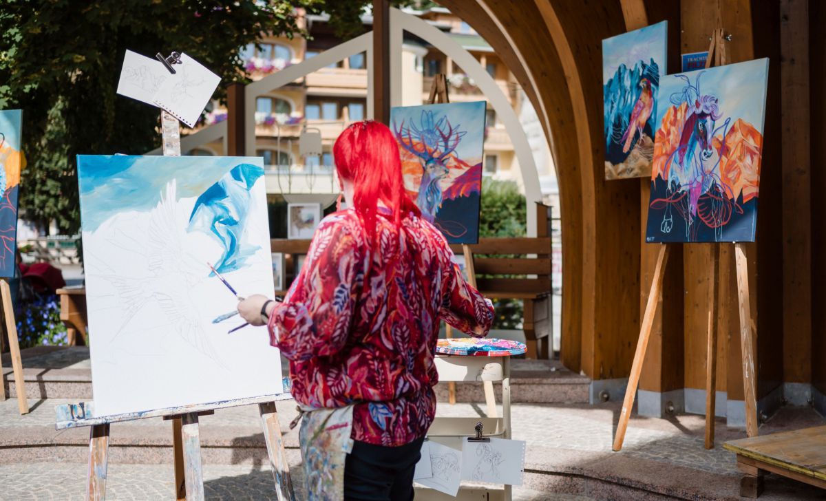 Frau mit langen roten Haaren malt auf einer Leinwand, dahinter mehrere fertige Bilder.