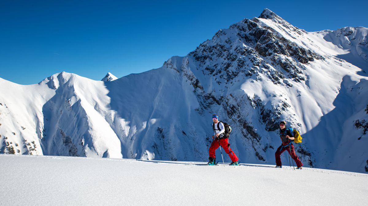 2 Skitourengeher, Tiefschnee. dahinter ein Berggipfel.