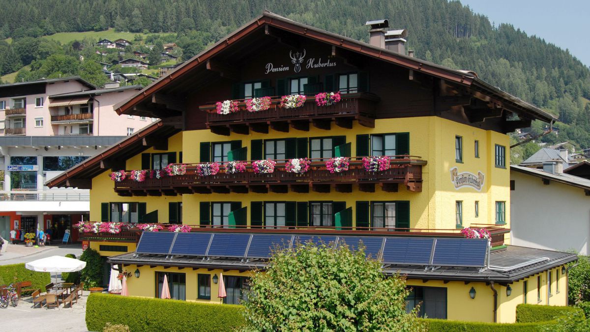 Aussenansicht der Pension: Gelbes Haus, dunkelbraube Balkongeländer mit roren und weißen Blumen, über dem vorragenden Speise´saal ist eine Solaranlage.