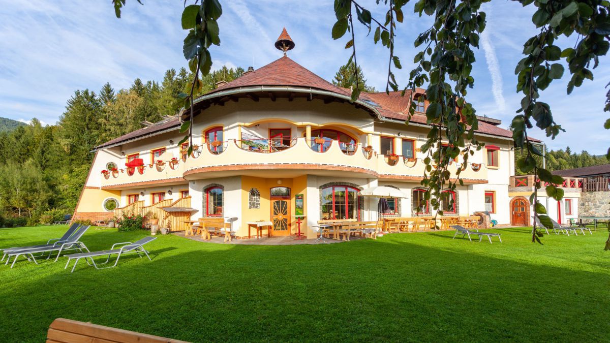 Außenaufnahme des Hotels im Sommer. In der Mitte win rundes Gebäude, links und rechts ein Ttrakt mit Zimmerm. Das Haus ist in gelb gehalten, Fenster und Blumen lassen es bunt erscheinen.