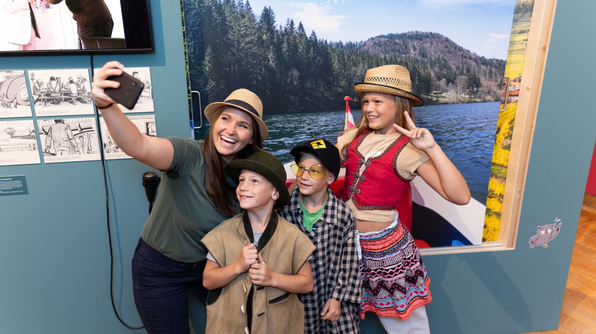 Frau mit drei Kindern macht ein Selfie vor einem Plakat mit dem Motiv eines Sees.