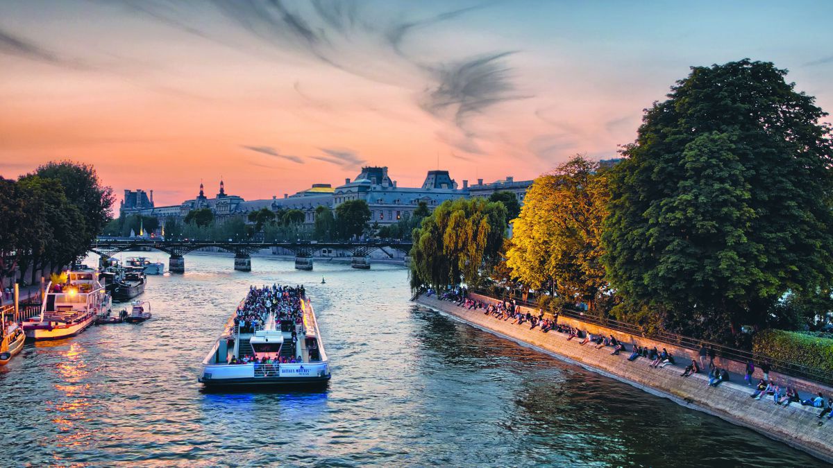 Blick auf die Seine in Paris: der rotgoldene Abendhimmel und die Lichter der Schiffe spiegeln sich im Fluss und im Laub der Bäume am Ufer wider.