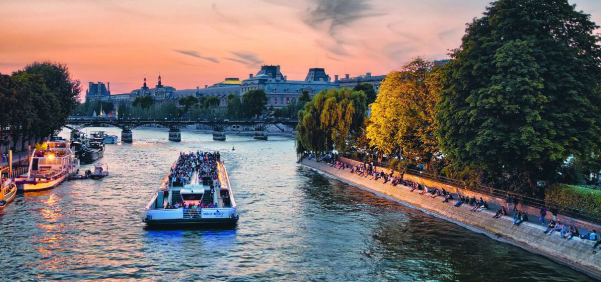 Blick auf die Seine in Paris: der rotgoldene Abendhimmel und die Lichter der Schiffe spiegeln sich im Fluss und im Laub der Bäume am Ufer wider.