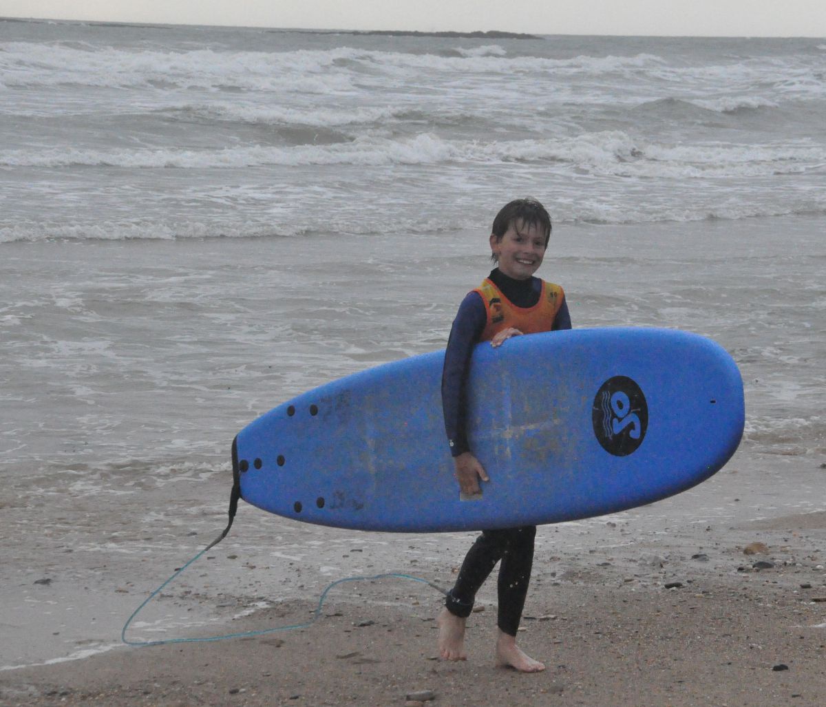 Ein Kind kommt gerade lächelnd aus dem Meer und trägt ein Surfbrett unter dem Arm.