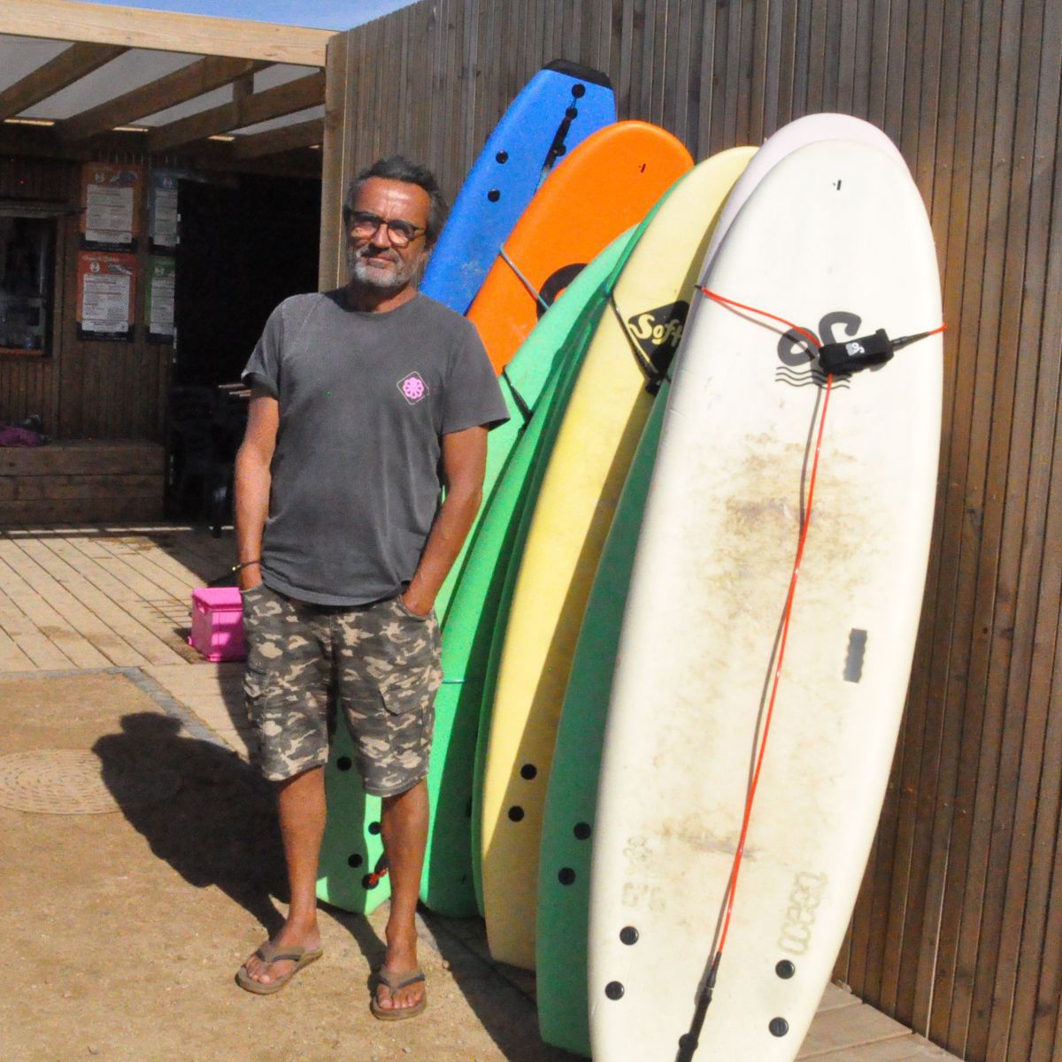 Momo posiert vor der Hütte der Surfschule neben einer Reihe bunter Surfbretter und blau-schwarzer Neoprenanzüge.