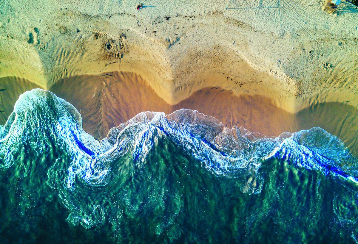 Meeresbrandung an sandiger Küste von oben fotografiert: Ein wellengeformtes Muster im Farbverlauf von dunklen und helleren Blautönen über Schaumweiß bis zu Ockergold und Sandfarben.