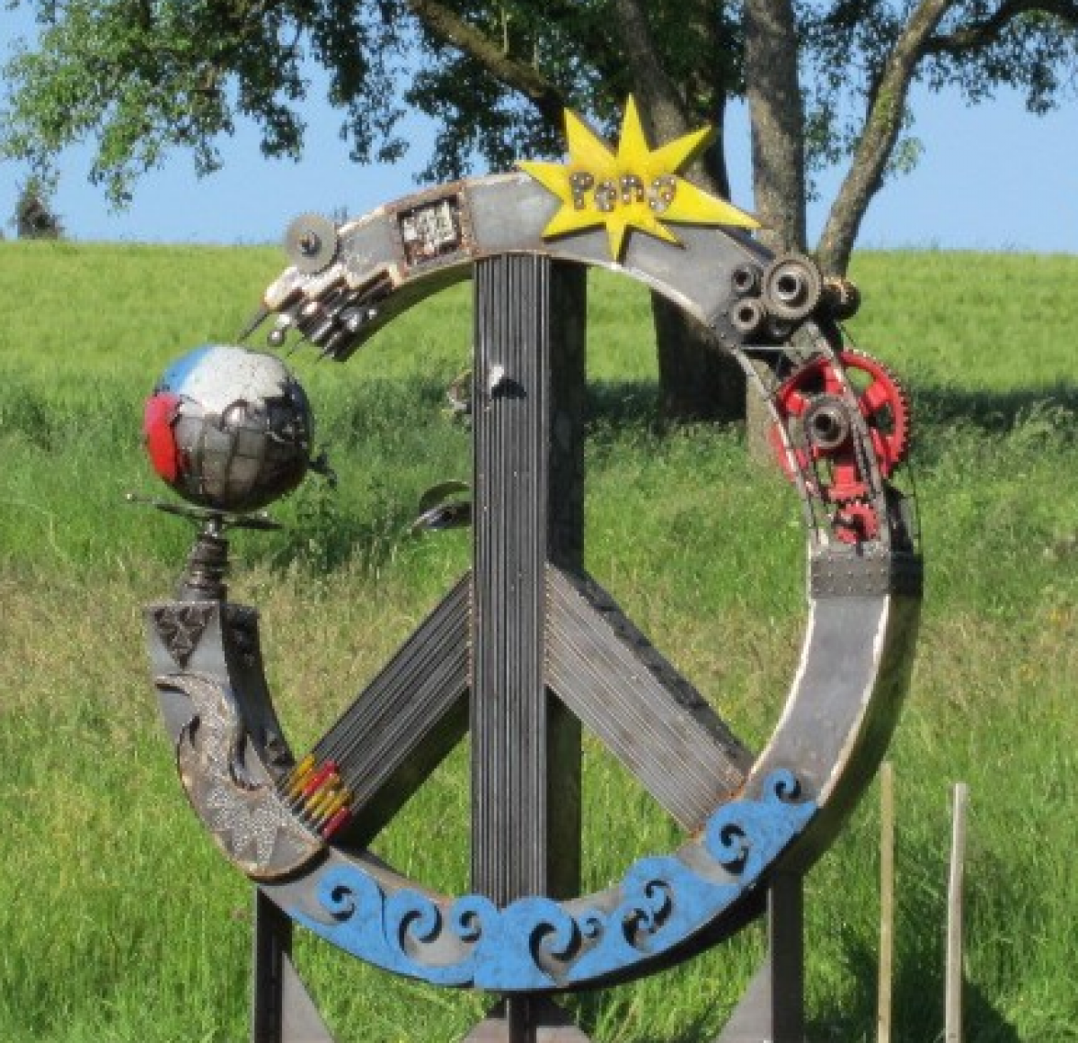 Eine Skulptur: Das Peace-Symbol aus Holz, darauf sind verschiedene andere Symbole angebracht: blaue Wasserwellen, Zahnräder, ein gelber Blitz mit der Aufschrift 