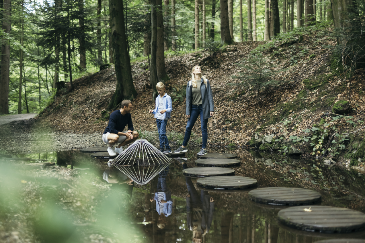 Ein Elternpaar mit Sohn im Schulalter gehen auf runden Holzinseln über die Wasserfläche am Erlebnisweg im Wald beim Ursprung des Flusses Trattnach.