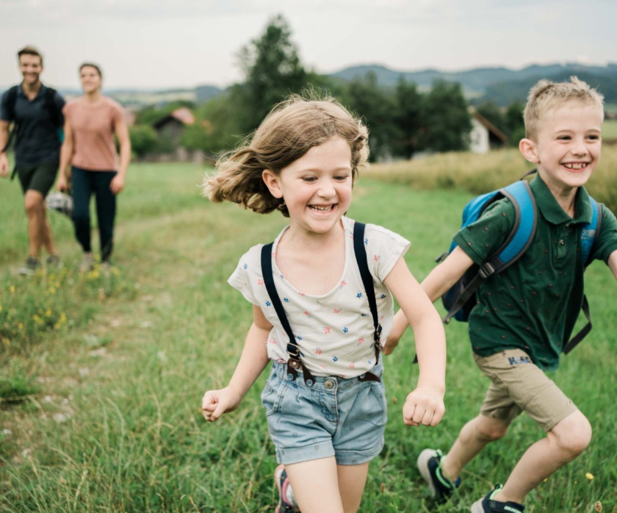 Ein Mädchen und ein Junge im Kindergartenalter laufen beim Wandern vom Weg ab auf eine Sommerwiese. Die Eltern gehen etwas weiter hinten. Die Familie ist sommerlich gekleidet, der Himmel ist leicht diesig, die Felder und Wiesen stehen hoch.